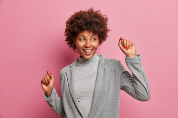 Wall Mural - Indoor shot of successful female winner dressed in grey formal clothing makes triumph dance smiles broadly looks aside poses against pink background. Executive woman worker rejoices success.