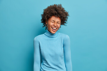 Portrait of overjoyed dark skinned Afro American woman laughs out loudly and keeps eyes closed has fun and giggles over something positive dressed in casual clothing isolated over blue background