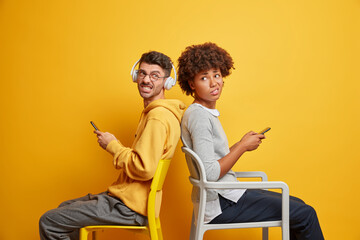 two mixed race friends wait for something sit back to each other on chairs smirk faces and have irri