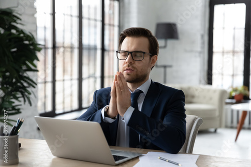 Hopeful young businessman sitting at workplace table with folded hands, praying God for good luck before online negotiations meeting or starting new project development on computer, feeling faithful.