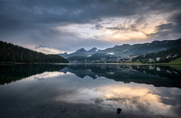 Wall Mural - crystal clear lake in a valley in switzerland in the evening with clouds and sun in front of mountains and a village
