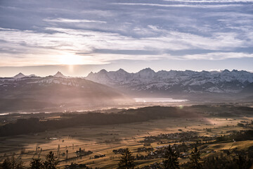 Wall Mural - overview swiss landscape with hills forests mountains in warm color during sunset or sunrise and epi