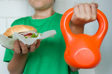 A boy in a green T-shirt holds a hamburger in one hand and an orange weight in the other. the concept of a healthy lifestyle.