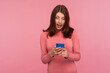 © khosrork - Excited brunette woman looking at smartphone display in her hands with big surprised eyes, receiving message about bonuses and discounts. Indoor studio shot isolated on pink background