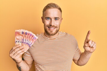 Wall Mural - Young caucasian man holding south african 50 rand banknotes smiling with an idea or question pointing finger with happy face, number one