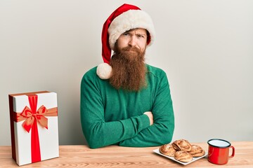 Canvas Print - Young irish redhead man wearing christmas sitting on the table with gift skeptic and nervous, disapproving expression on face with crossed arms. negative person.