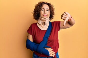 Wall Mural - Beautiful middle age mature woman wearing cervical collar and arm on sling with angry face, negative sign showing dislike with thumbs down, rejection concept