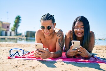 Young latin couple using smartphone lying on the sand at the beach.