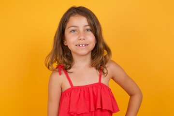 Studio shot of cheerful young Caucasian girl standing against yellow background keeps hand on hip, smiles broadly.