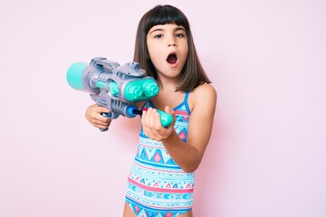 Wall Mural - Young little girl with bang wearing swimsuit and watergun in shock face, looking skeptical and sarcastic, surprised with open mouth