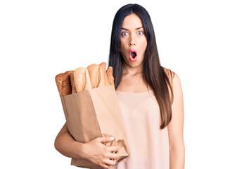 Poster - Young beautiful caucasian woman holding paper bag with bread scared and amazed with open mouth for surprise, disbelief face