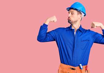 Young handsome man wearing worker uniform and hardhat showing arms muscles smiling proud. fitness concept.