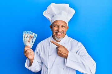 Poster - Middle age grey-haired man wearing professional cook uniform holding swiss franc banknotes smiling happy pointing with hand and finger