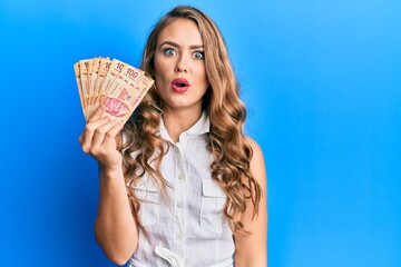 Sticker - Young blonde girl holding mexican pesos scared and amazed with open mouth for surprise, disbelief face