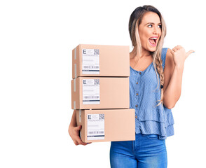 Sticker - Young beautiful blonde woman holding delivery boxes pointing thumb up to the side smiling happy with open mouth