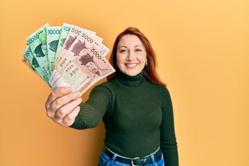 Wall Mural - Beautiful redhead woman holding south korean won banknotes looking positive and happy standing and smiling with a confident smile showing teeth