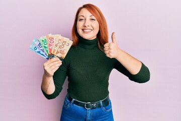 Sticker - Beautiful redhead woman holding canadian dollars smiling happy and positive, thumb up doing excellent and approval sign