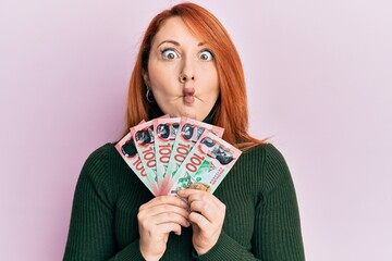 Poster - Beautiful redhead woman holding 100 new zealand dollars banknote making fish face with mouth and squinting eyes, crazy and comical.