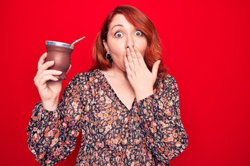 Sticker - Young beautiful redhead woman drinking mate infusion beverage over isolated red background covering mouth with hand, shocked and afraid for mistake. Surprised expression