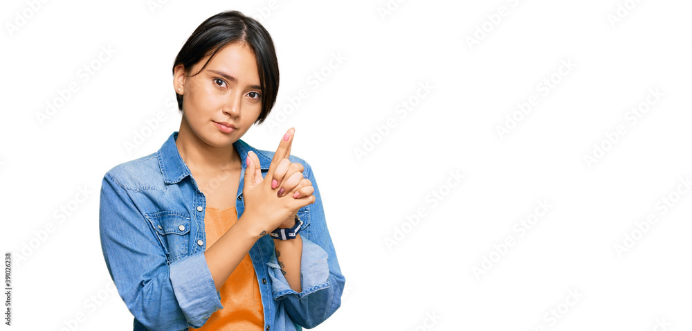 Young beautiful hispanic woman with short hair wearing casual denim jacket holding symbolic gun with hand gesture, playing killing shooting weapons, angry face
