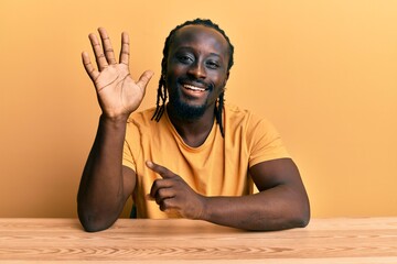 Poster - Handsome young black man wearing casual clothes sitting on the table showing and pointing up with fingers number five while smiling confident and happy.