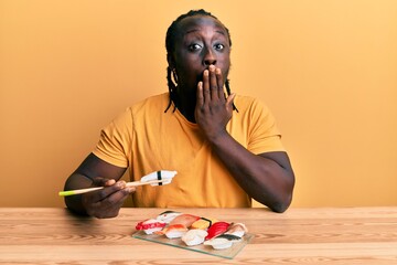 Canvas Print - Handsome young black man eating sushi sitting on the table covering mouth with hand, shocked and afraid for mistake. surprised expression