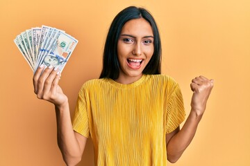 Sticker - Young brunette woman holding dollars pointing thumb up to the side smiling happy with open mouth