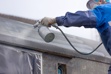 Industrial work. Priming of metal products from the compressor gun. A worker in overalls and a protective mask paints the body of a truck trailer or a metal car.