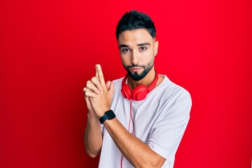 Canvas Print - Young man with beard listening to music using headphones holding symbolic gun with hand gesture, playing killing shooting weapons, angry face