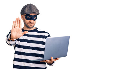 Sticker - Young handsome man wearing burglar mask using laptop with open hand doing stop sign with serious and confident expression, defense gesture