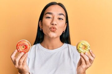 Sticker - Young asian woman holding tasty colorful doughnuts looking at the camera blowing a kiss being lovely and sexy. love expression.