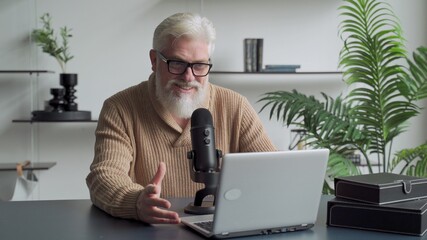 Elderly man with a gray beard video blogger speaking in microphone. Aged broadcaster lives in a studio