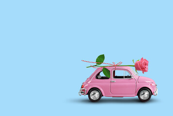  Pink toy car delivering pink rose flower on a blue background. Flowers delivery, women day, valentine day.