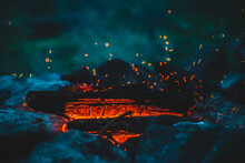 Vivid Smoldered Firewoods Burned In Fire Close-up. Atmospheric Background With Orange Flame Of Campfire And Blue Smoke. Warm Full Frame Image Of Bonfire. Glowing Embers In Air. Bright Sparks In Bokeh