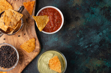 Wall Mural - Appetizing corn tortilla chips on dark blue marble background. Salsa, cheese dip and avocado souse for taste. Unhealthy food. Traditional mexican dish. View from above. Concept of food process.