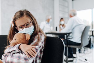 Wall Mural - Cute little girl with protective face mask and toy in hands waiting her grandfather in hallway while he is at doctor.