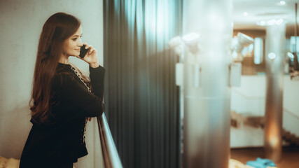 Charming mature woman entrepreneur with long hair is leaning on a chrome balcony fence and talking on the phone while standing in a large business office chillout area; a copy space place on the right