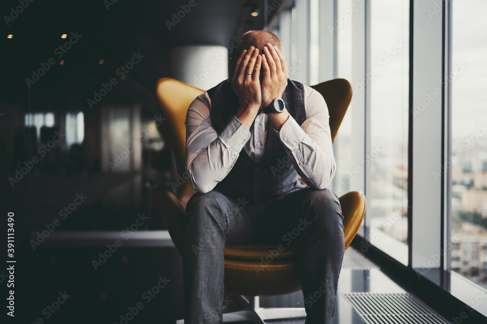 A man entrepreneur is in sorrow because of his dismissal; a businessman on an orange armchair near an office window covered his face with his hands after learning about the bankruptcy of the company - obrazy, fototapety, plakaty 