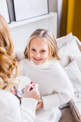 Wall Mural - Mother and daughter in light sweaters are sitting on the sofa, the room is decorated for the new year and Christmas, hugs, conversations, smiles, kisses, waiting for a gift and magic