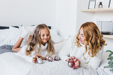 Wall Mural - Mom and daughter blonde hair and white sweaters lie on the bed, light up, say toys for Christmas and new year tree, Christmas tree decoration