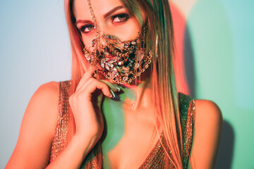 Wall Mural - Quarantine style. Neon light portrait. Fashion trend 2021. Mysterious woman in glamour chain gem face mask matching gold party outfit posing in pastel green blue glow.