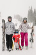 © Serhii - Skiers stand mountain top with skis in hands. Skiing concept. Ski resort. Family enjoying winter vacation on snow in cold snowy day. Happy dad and mom with child daughter having fun and hugging.