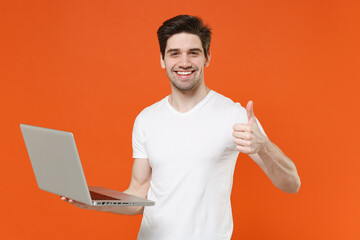 Smiling cheerful funny young man in basic blank white t-shirt standing working on laptop pc computer showing thumb up looking camera isolated on bright orange colour wall background, studio portrait.