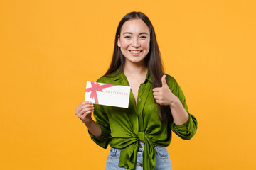 Wall Mural - Smiling funny young brunette asian woman wearing basic green shirt standing hold in hand gift certificate showing thumb up looking camera isolated on bright yellow colour background, studio portrait.