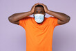 © ViDi Studio - Worried young african american man wearing basic casual orange t-shirt sterile face mask to safe from coronavirus virus covid-19 put hands on head isolated on pastel violet background studio portrait.