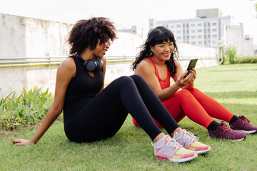 Diverse group of two female fitness friends talking and using a mobile phone in the park. Young women resting and smiling outdoors