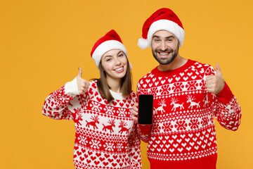 Wall Mural - Smiling young Santa couple friends man woman in sweater, Christmas hat hold mobile phone with blank empty screen showing thumbs up isolated on yellow background. Happy New Year celebration concept.