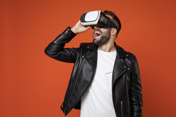 Wall Mural - Excited cheerful young bearded man 20s wearing casual basic white t-shirt, black leather jacket standing watching in vr headset gadget isolated on bright orange colour background studio portrait.