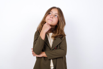 Wall Mural - Portrait of thoughtful Beautiful little girl standing against white background,  keeps hand under chin, looks away trying to remember something or listens something with interest. Youth concept.