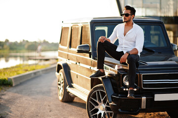 Solid asian man in white shirt and sunglasses posed near black mafia suv car.